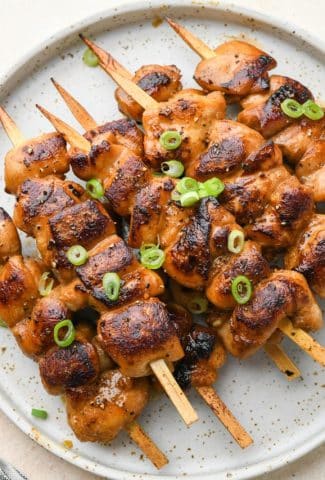 Cooked garlic ginger chicken skewers piled on a plate and topped with thinly sliced green onions.