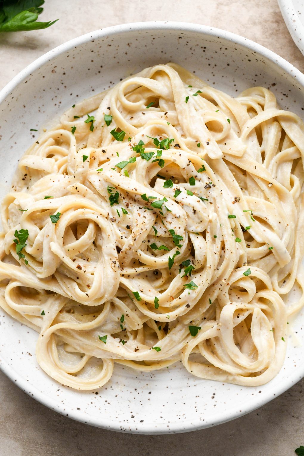 A bowl of dairy free fettuccine alfredo on a light brown colored background. Topped with black pepper and fresh herbs.