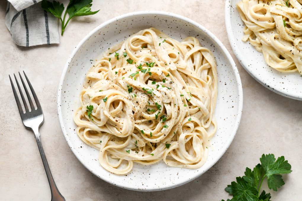 Two side by side bowls of dairy free fettuccine alfredo on a light brown colored background. Topped with black pepper and fresh herbs.