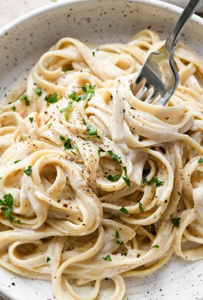 A bowl of vegan fettuccine alfredo topped with fresh parsley and black pepper with a fork twirling some noodles.