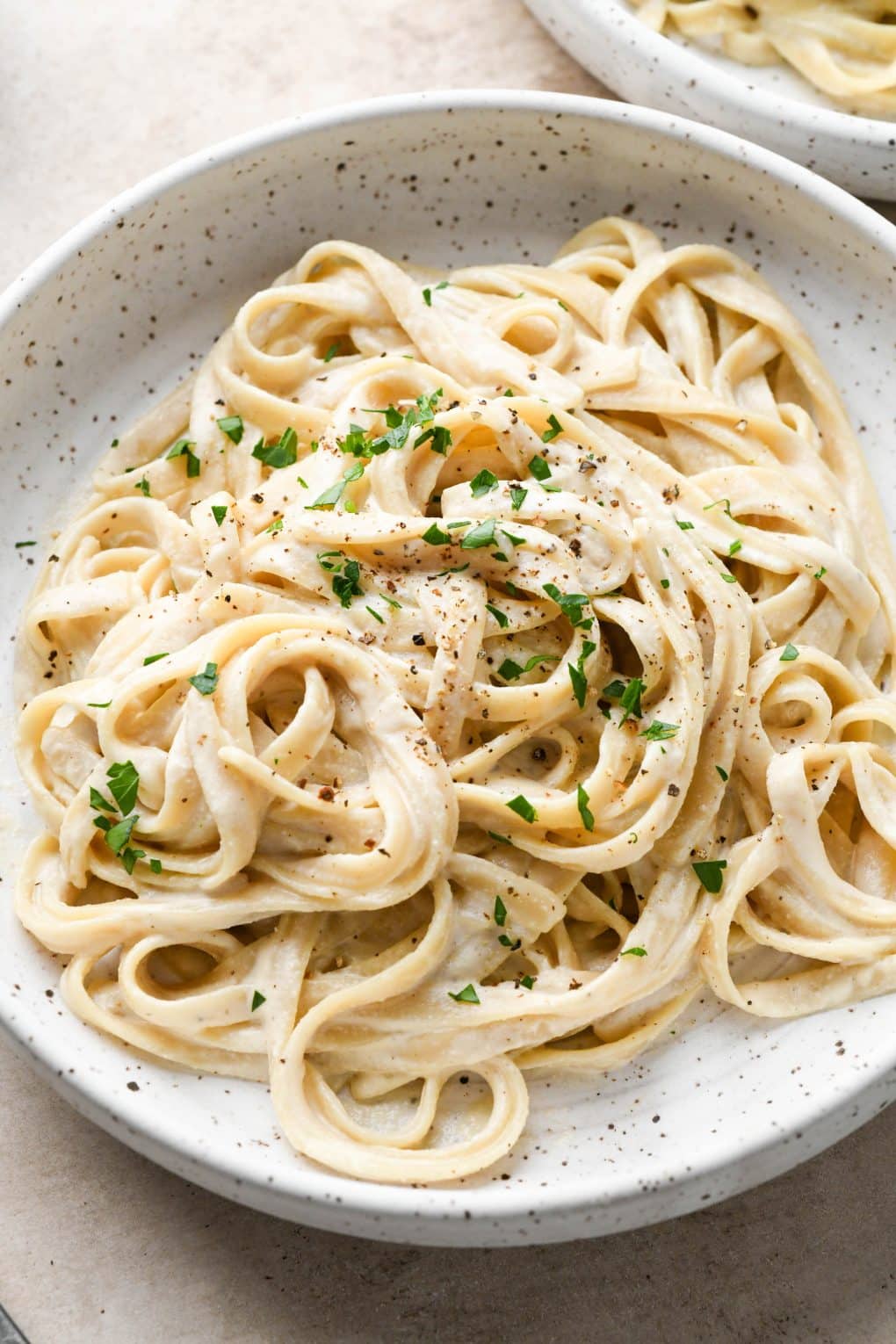 A bowl of dairy free fettuccine alfredo on a light brown colored background. Topped with black pepper and fresh herbs.