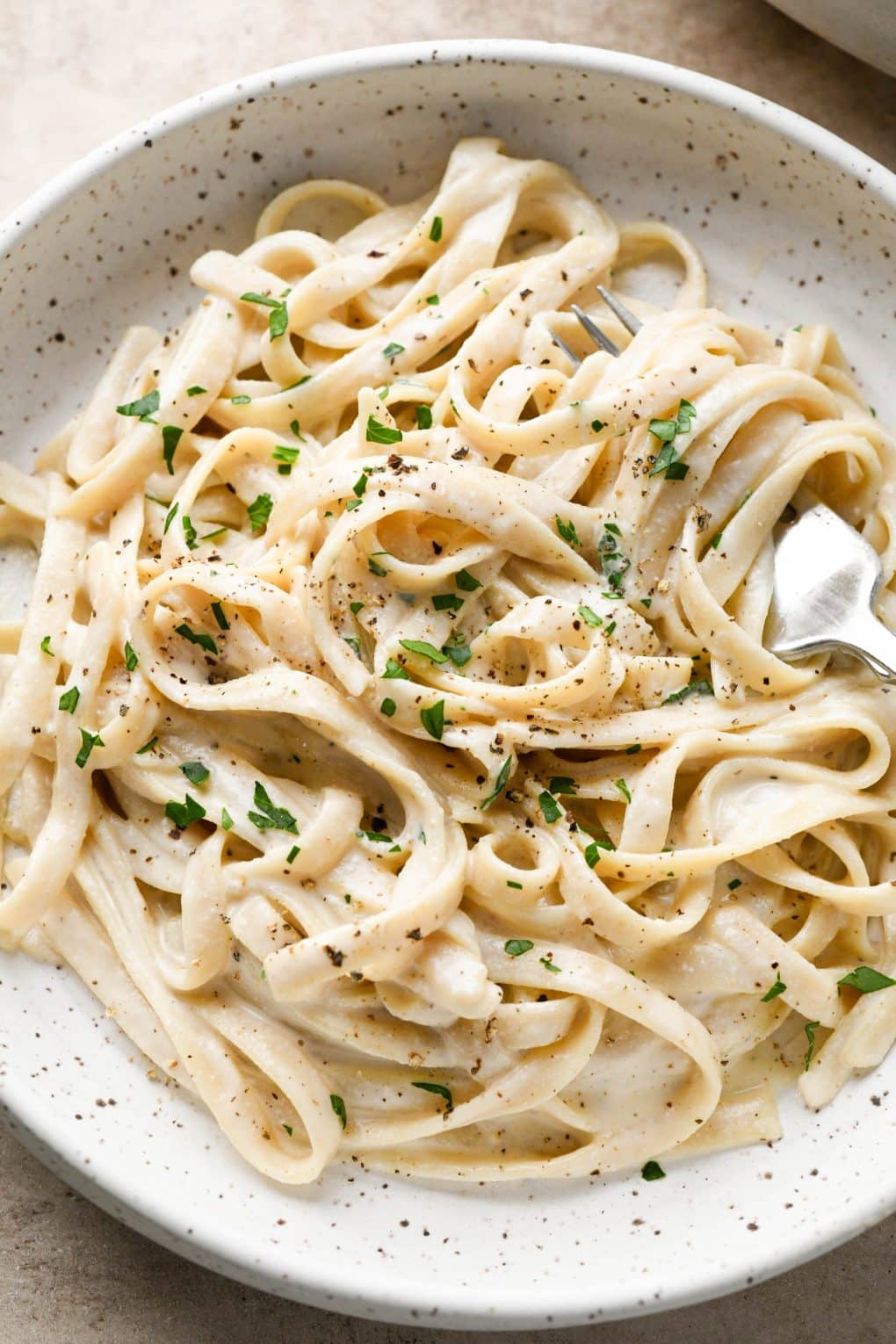 A bowl of vegan fettuccine alfredo topped with fresh parsley and black pepper with a fork twirling some noodles.