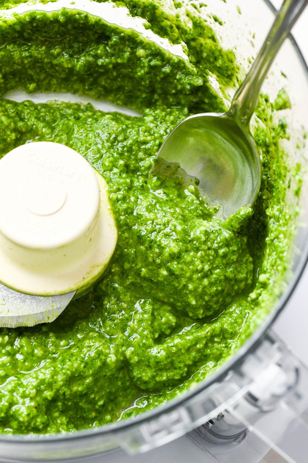 Homemade basil pesto in a food processor container with a spoon dipping into the sauce.