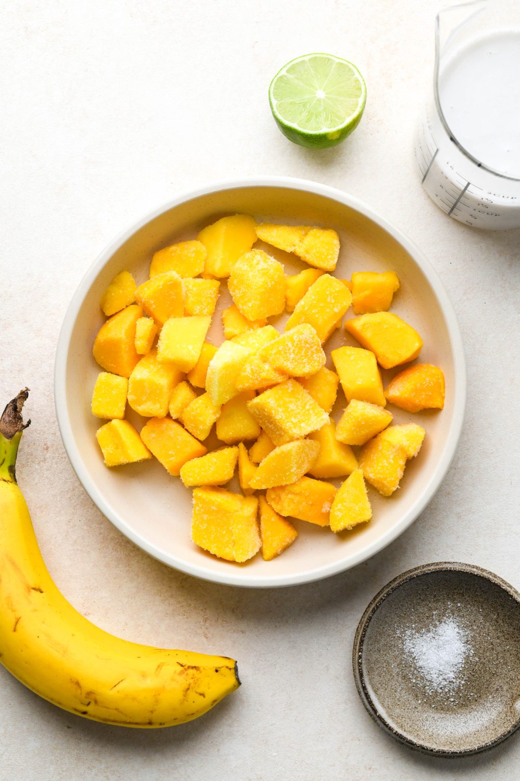 Ingredients for simple mango smoothies in various ceramics, on a cream colored background.