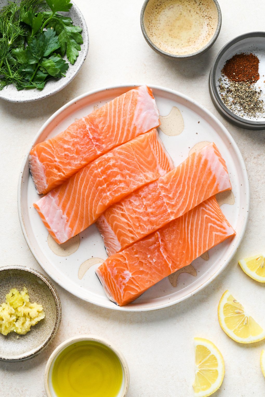 Ingredients for baked salmon on various sized ceramic plates and bowls, on a light cream colored background.