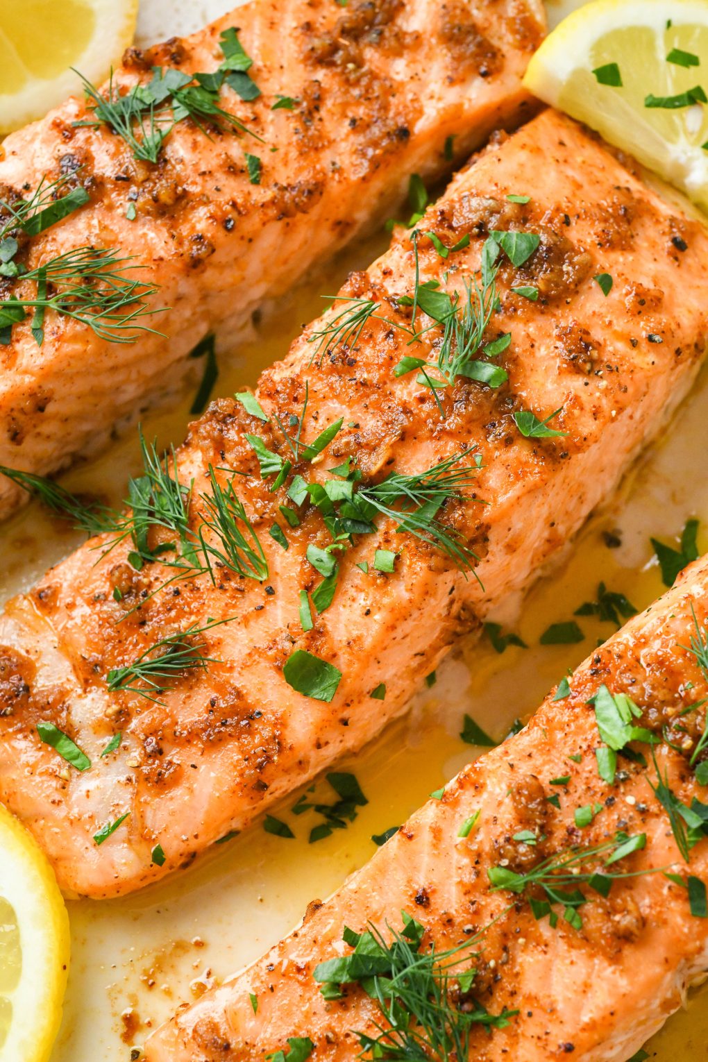 Oven baked salmon in a baking dish topped with fresh parsley, dill, and lemon wedges.