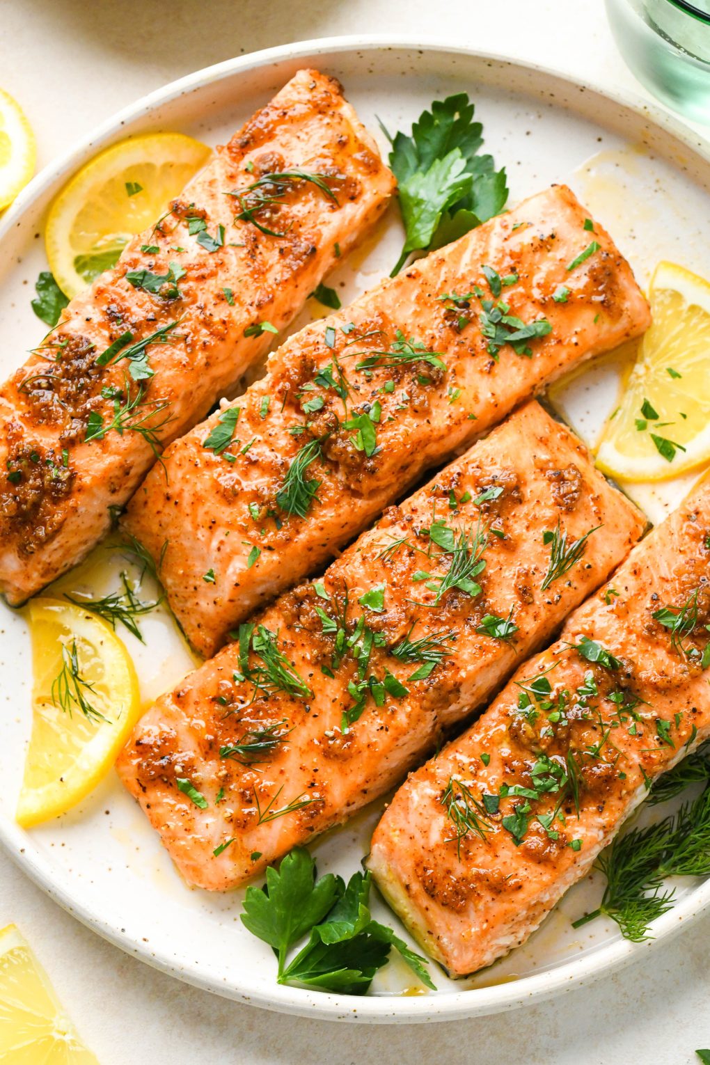 Oven baked salmon fillets on a cream colored ceramic plate, topped with fresh herbs and lemon wedges.