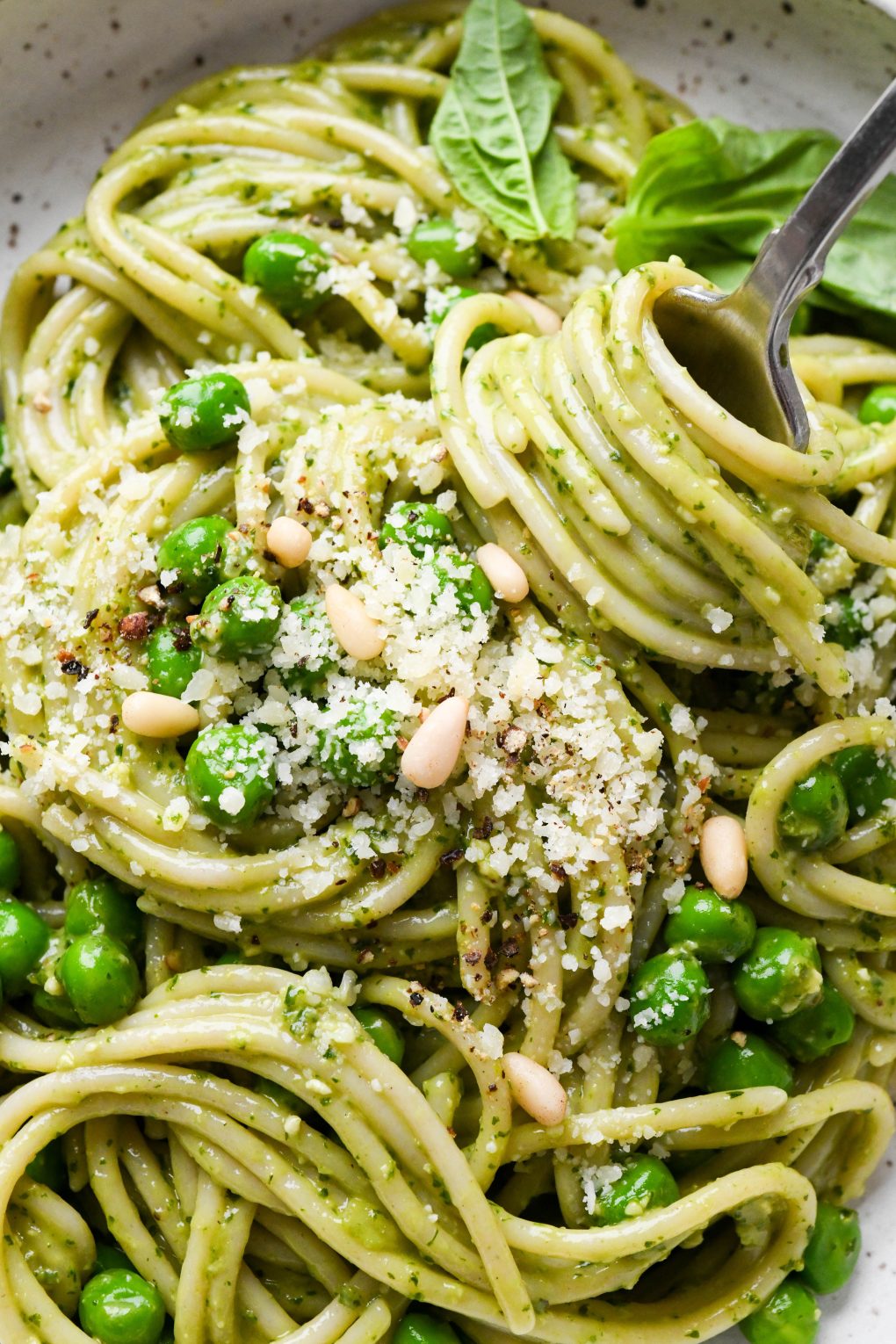 Pesto pasta made with spaghetti in a speckled ceramic bowl with a fork twirling a bite, topped with pine nuts and grated parmesan.