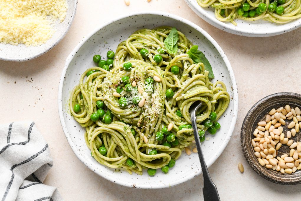 Pesto pasta made with spaghetti twirled in a speckled ceramic bowl and topped with pine nuts and grated parmesan. Next to a small dish of parmesan cheese, pine nuts, and a second bowl of pasta.