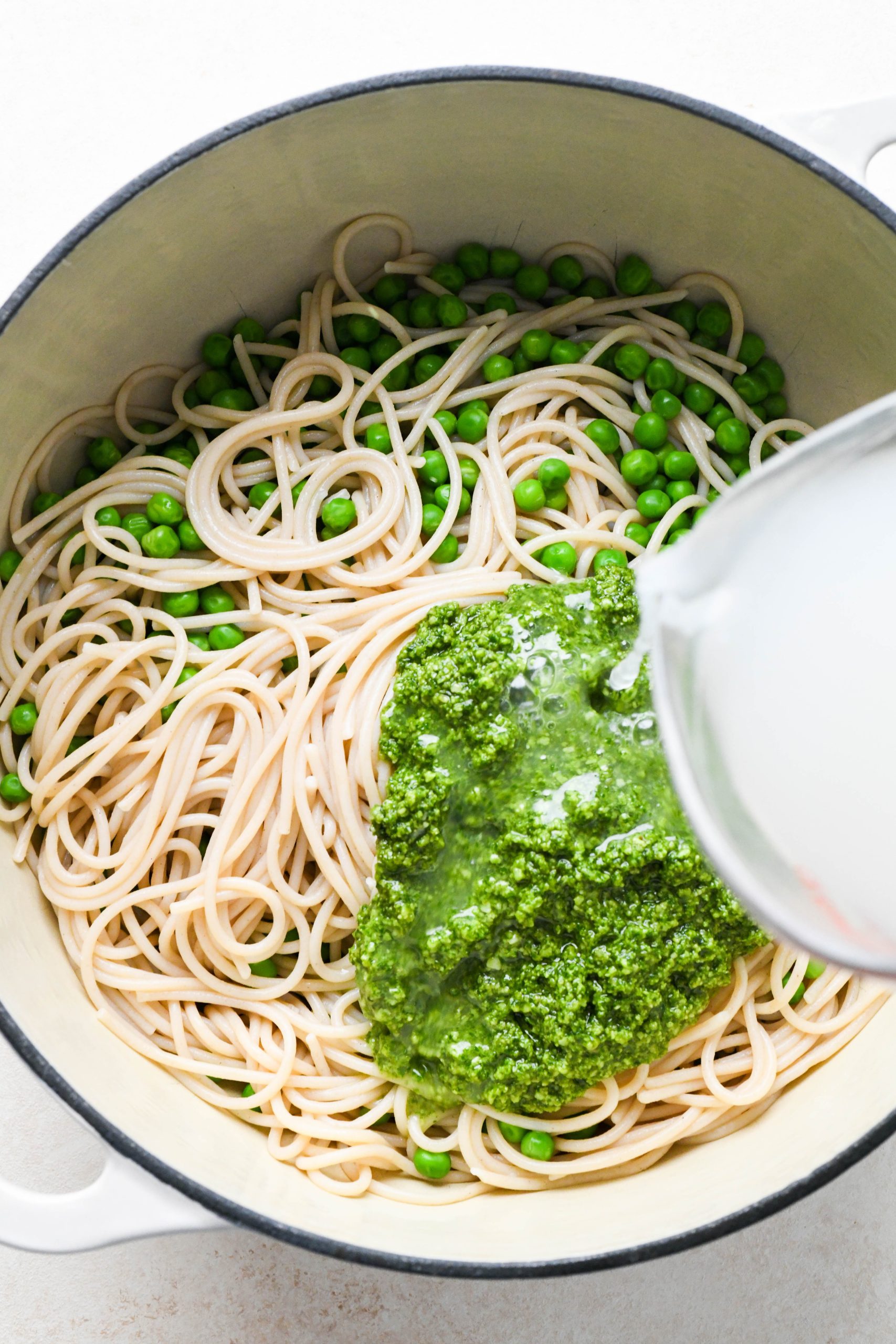 How to make easy pesto pasta: Pasta and peas in pot with pesto on top, a hand pouring pasta cooking water over the pesto.