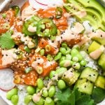 Homemade poke bowl topped with edamame, cucumber salad, radishes, green onions, thinly sliced jalapeño, avocado, cilantro, sesame seeds, and spicy mayo in a shallow speckled bowl.