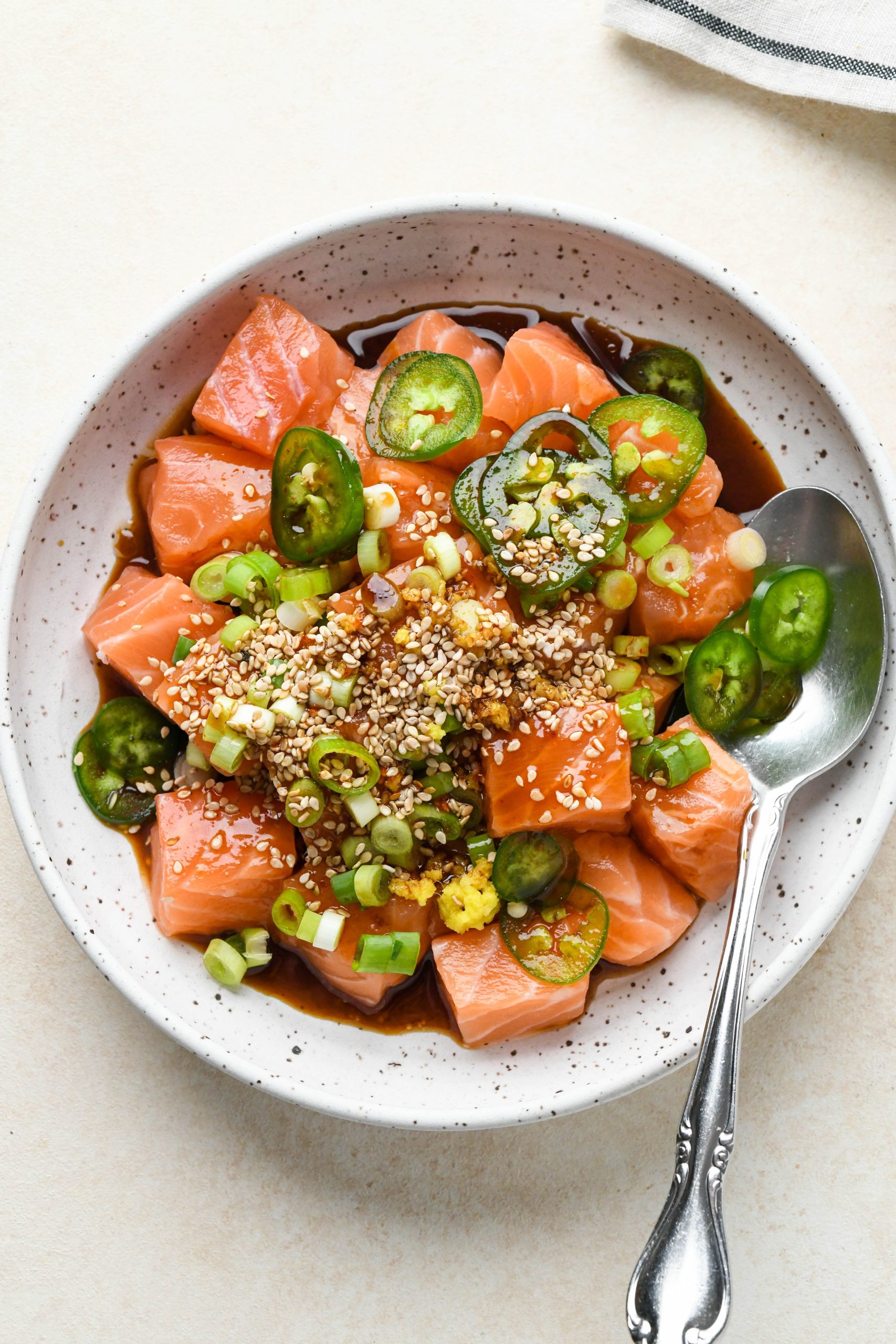 How to make poke bowls: Raw salmon with jalapeño, grated ginger, green onions, and sesame seeds in a shallow bowl topped with poke sauce, before mixing.