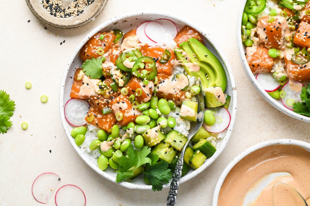 Two side by side homemade poke bowls topped with edamame, cucumber salad, radishes, green onions, thinly sliced jalapeño, avocado, cilantro, sesame seeds, and spicy mayo in a shallow speckled bowl.