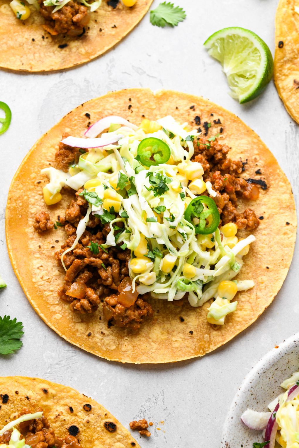 A single ground pork taco laid out on a concrete surface, topped with a creamy cabbage and corn slaw, with jalapeño, cilantro, and lime wedges.