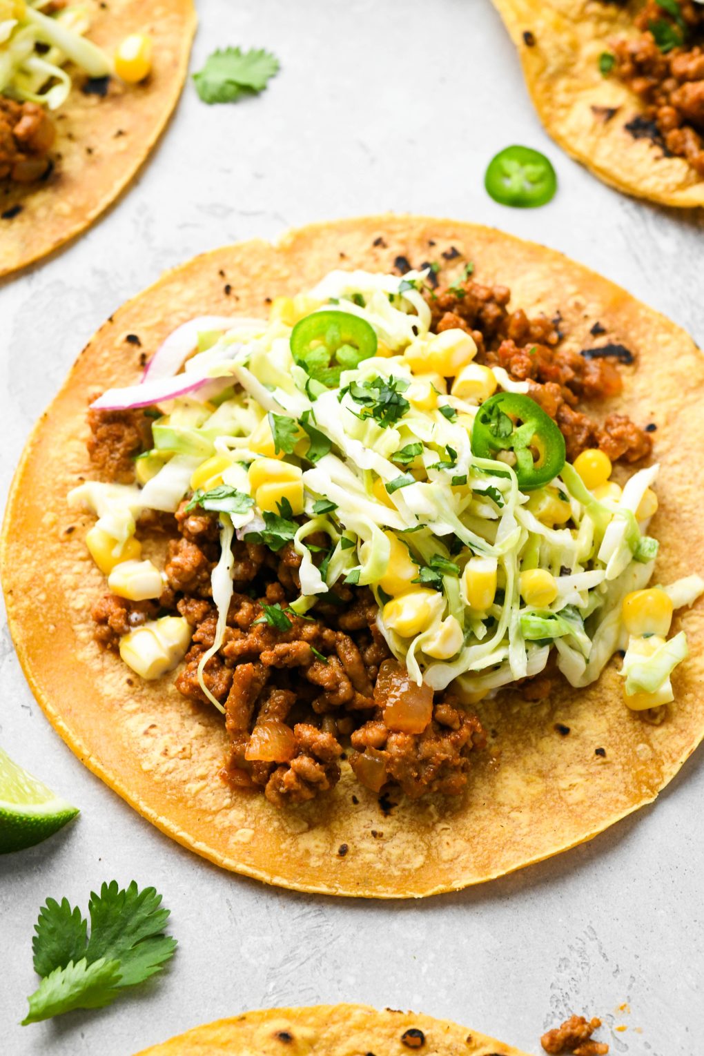 Detailed side angle view of a single ground pork taco laid out on a concrete surface, topped with a creamy cabbage and corn slaw, with jalapeño, cilantro, and lime wedges.