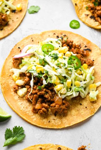 Detailed side angle view of a single ground pork taco laid out on a concrete surface, topped with a creamy cabbage and corn slaw, with jalapeño, cilantro, and lime wedges.