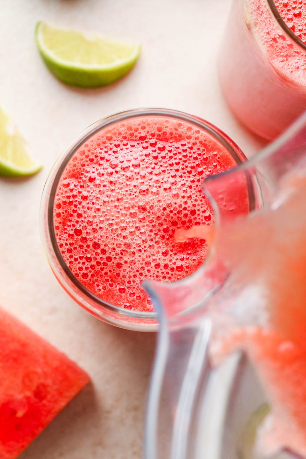 A blender container pouring watermelon juice into a cup on a light cream background, surrounded by lime wedges and cut watermelon slices.