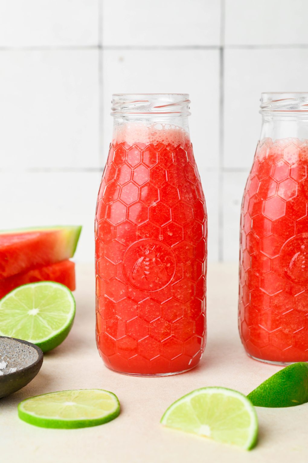 Bright pink watermelon juice in two small textured glass jars, next to a stack of cut up watermelon and some lime wedges.