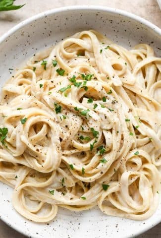 A bowl of dairy free fettuccine alfredo on a light brown colored background. Topped with black pepper and fresh herbs.