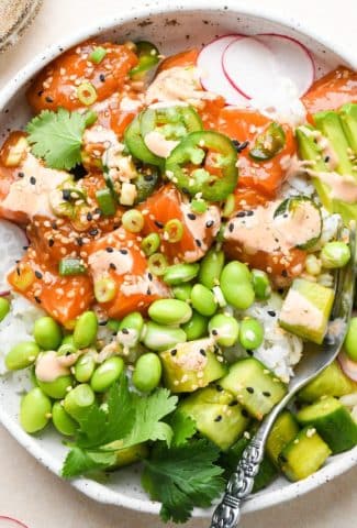 Homemade poke bowl topped with edamame, cucumber salad, radishes, green onions, thinly sliced jalapeño, avocado, cilantro, sesame seeds, and spicy mayo in a shallow speckled bowl.