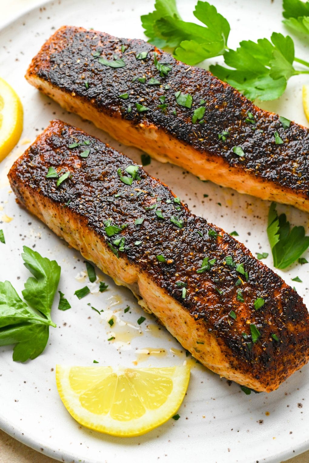 Two blackened salmon filets on a white speckled plate, surrounded by fresh herbs and lemon wedges.