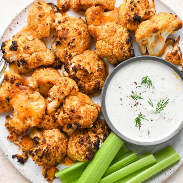 Buffalo cauliflower florets on a large round speckled plate next to a small bowl of ranch and celery sticks.