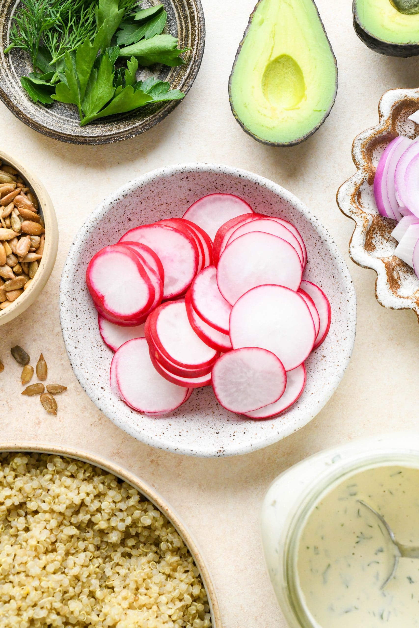 Ingredients for buffalo roasted cauliflower bowls - radishes in a small dish, sliced avocado, sunflower seeds, quinoa, and red onion in various ceramics.