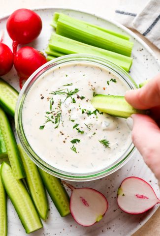 Vegan ranch dressing in a small glass jar with a hand dipping a cucumber slice surrounded by fresh cut veggies.