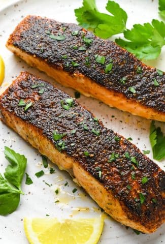 Two blackened salmon filets on a white speckled plate, surrounded by fresh herbs and lemon wedges.