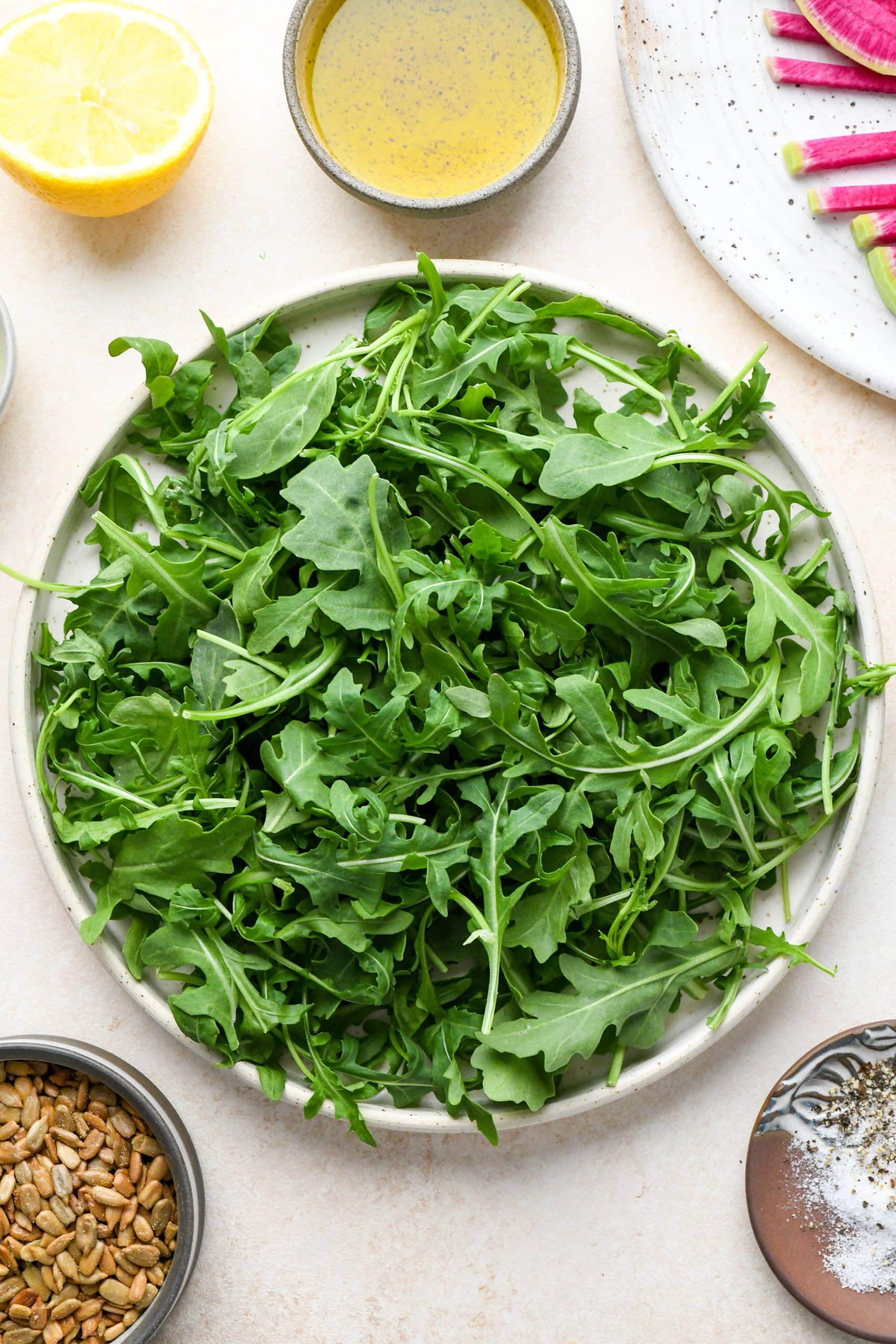 Ingredients for Simple Arugula Salad: Arugula, sunflower seeds, radishes, and dressing ingredients on a light cream colored background.