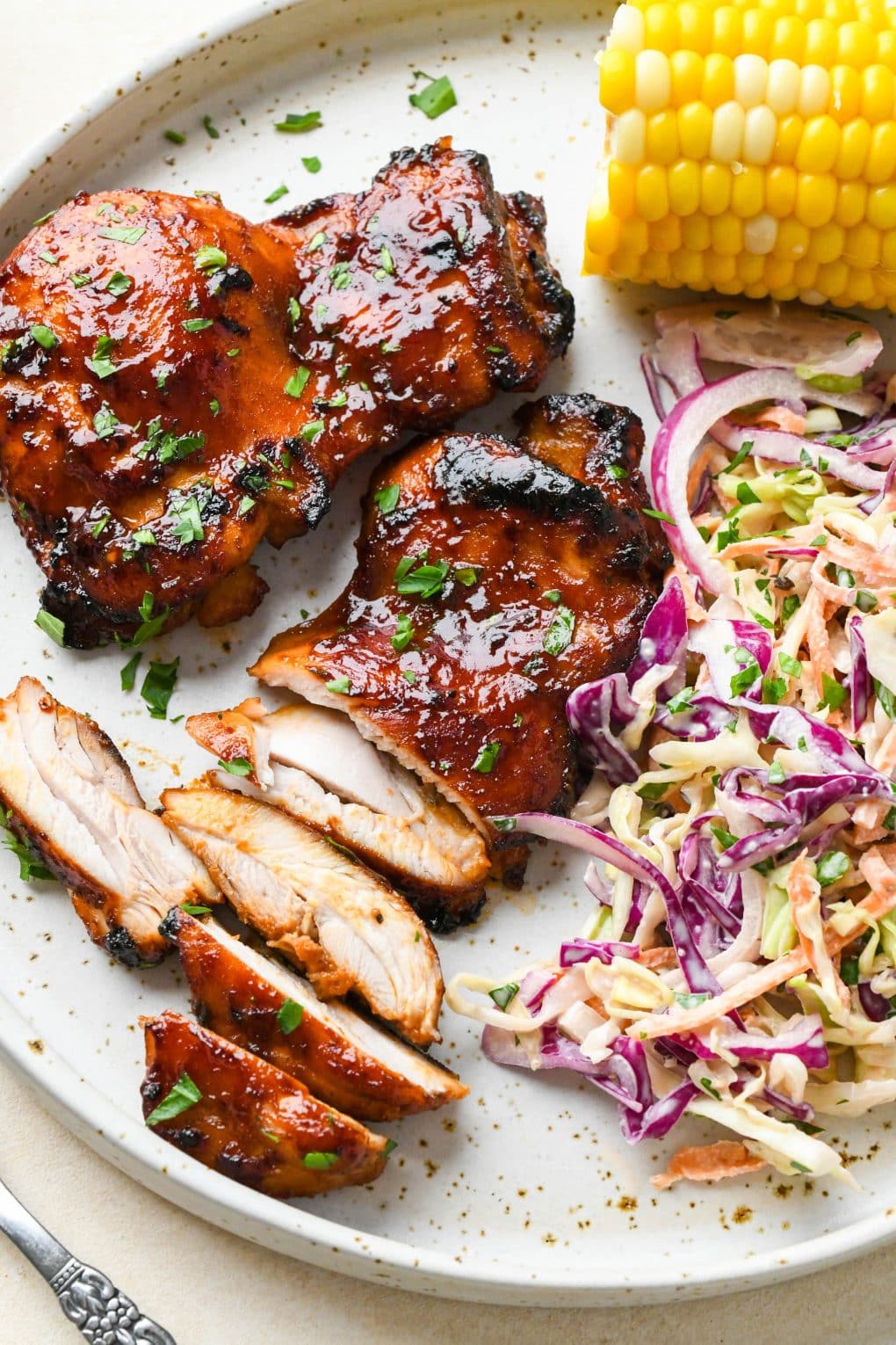 A large white ceramic plate of boneless skinless bbq chicken thighs next to corn on the cob and creamy coleslaw.
