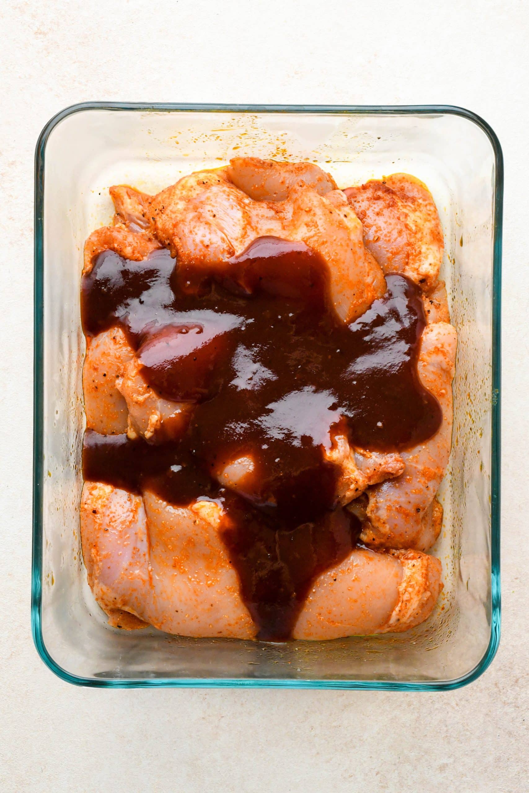 How to make BBQ Chicken Thighs in the Oven: Marinated chicken thighs in a glass container topped with bbq sauce.