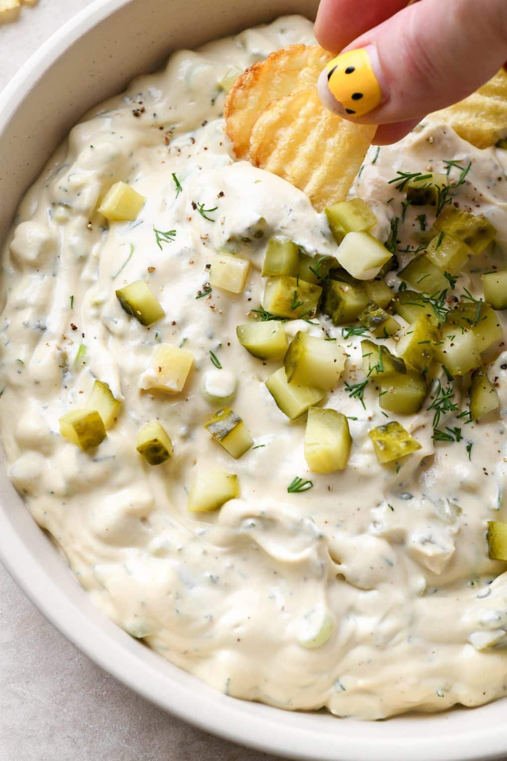 A hand dipping a ruffled potato chip into dairy free dill pickle dip.