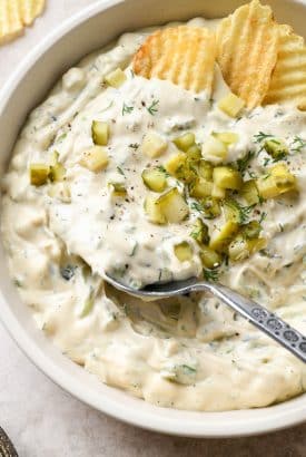 Large shallow ceramic bowl filled with super creamy dairy free dill pickle dipped. Topped with diced pickles and a large spoon is dipping into the dip.