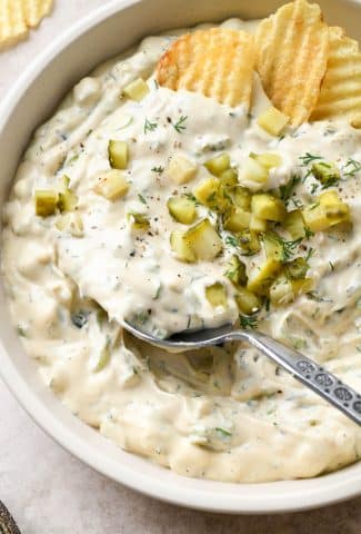 Large shallow ceramic bowl filled with super creamy dairy free dill pickle dipped. Topped with diced pickles and a large spoon is dipping into the dip.