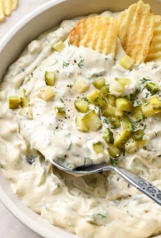 Large shallow ceramic bowl filled with super creamy dairy free dill pickle dipped. Topped with diced pickles and a large spoon is dipping into the dip.