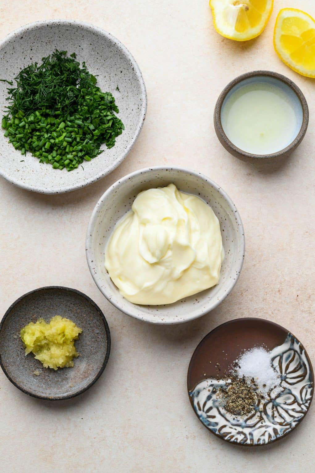 Ingredients for garlic herb aioli in a variety of ceramic dishes on a cream colored background.