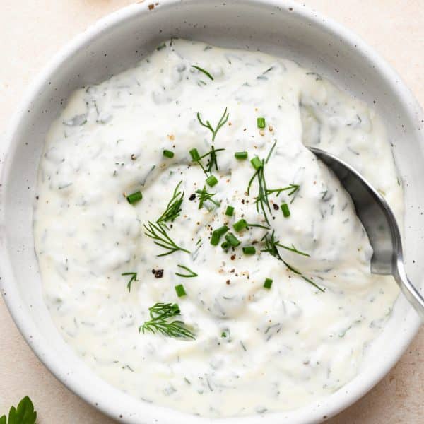 A small shallow ceramic bowl of herb flecked garlic aioli, topped with extra fresh herbs with a spoon dipping into the aioli.
