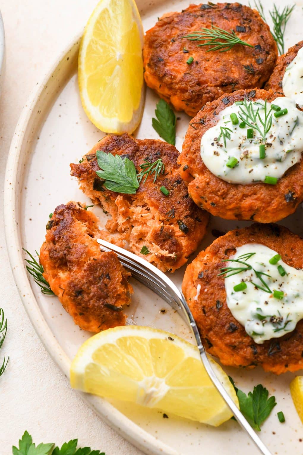 A fork cutting into a Whole30 salmon cake, on a platter of patties topped with garlic herb aioli and fresh herbs. 