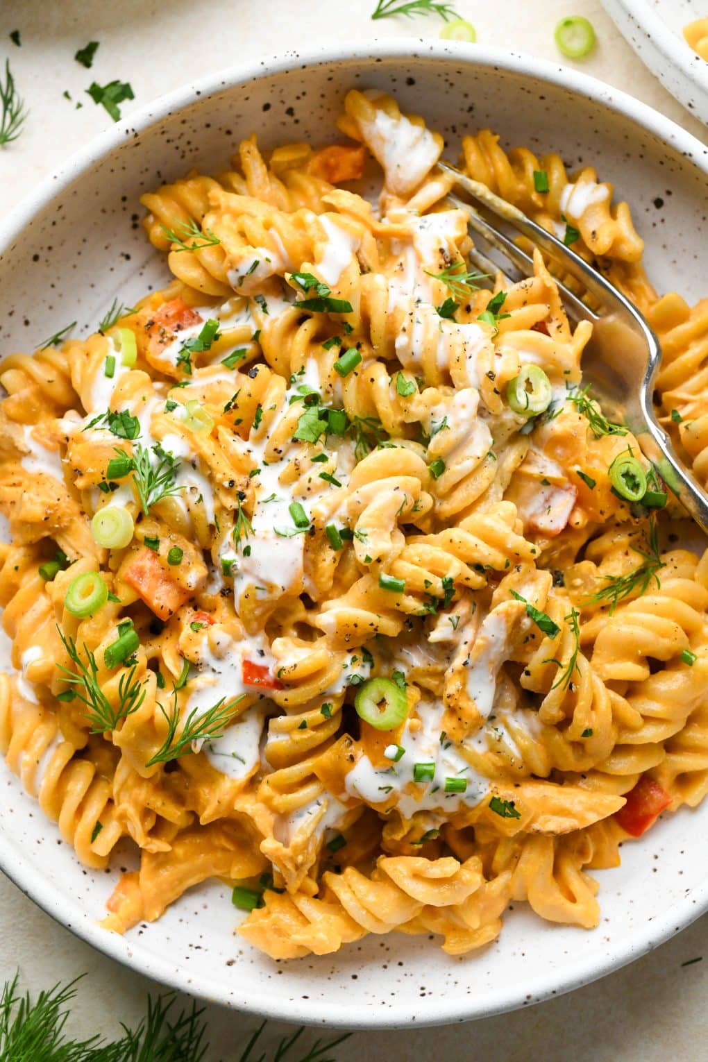 A shallow speckled bowl of creamy dairy free buffalo chicken pasta topped with fresh herbs, dairy free ranch, and a fork angled into the bowl.