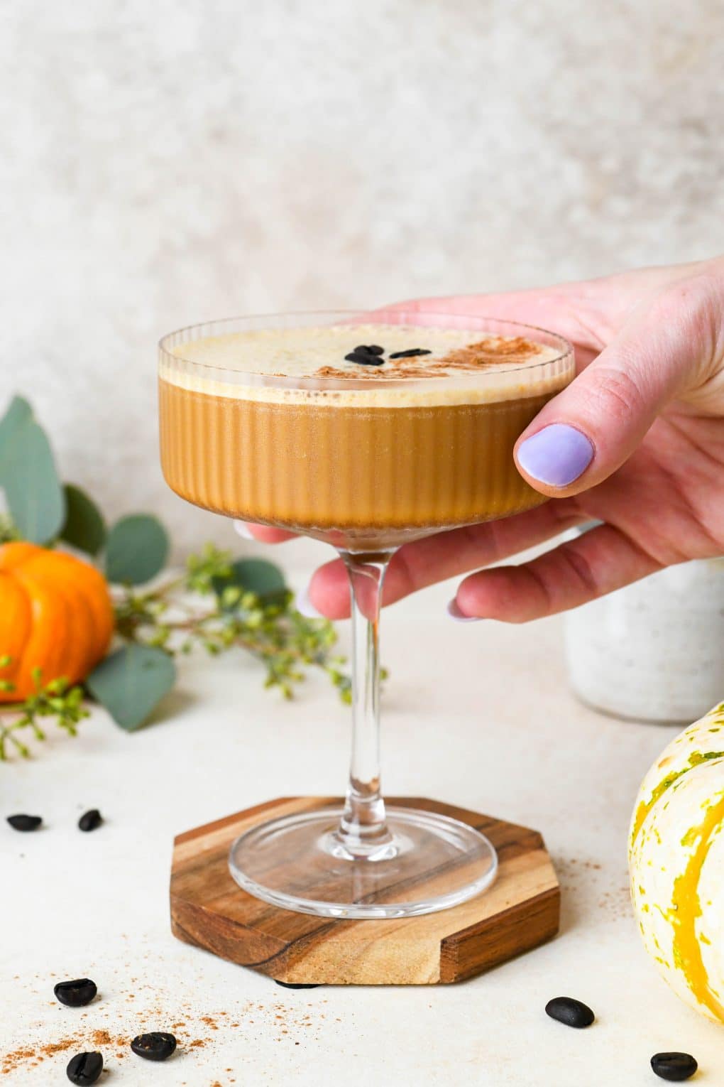 A pumpkin spice espresso martini on a coaster, with a hand reaching in to the image to pick it up. 