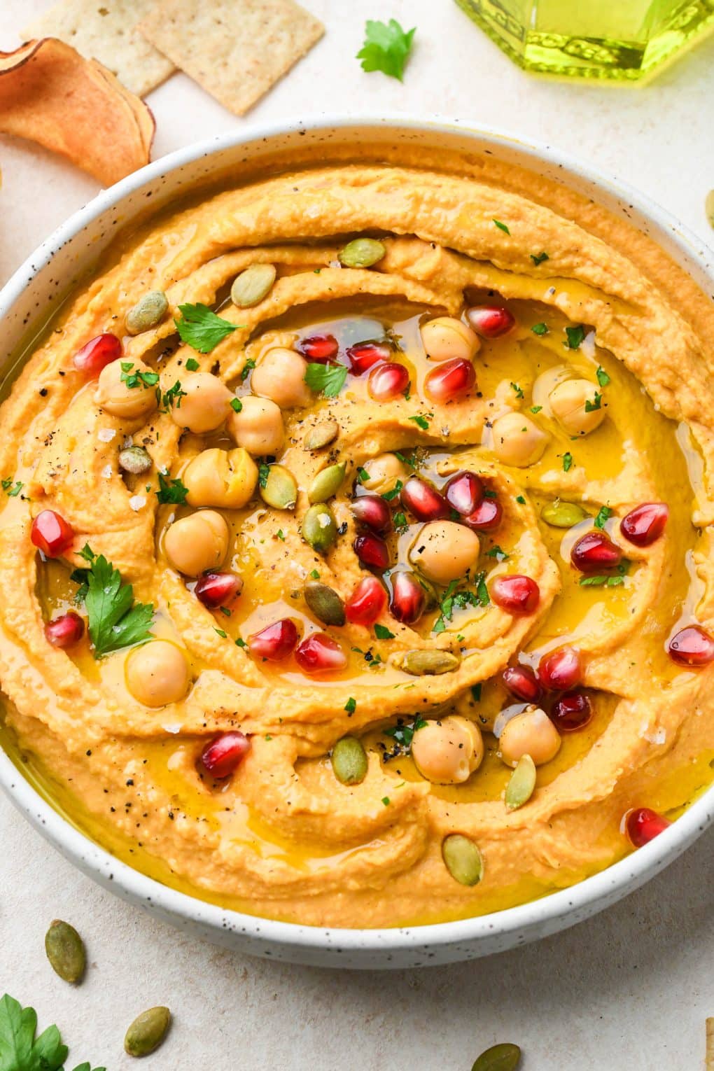 Close up image of a bowl of creamy sweet potato hummus topped with all the things - pomegranate, fresh parsley, olive oil, chickpeas, and flaky salt and pepper.