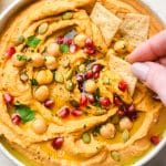 A shallow bowl of swirled sweet potato hummus topped with fresh herbs, pumpkin seeds, olive oil, and pomegranate with a hand dipping a small cracker into the dip.