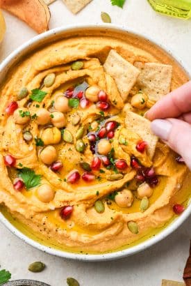 A shallow bowl of swirled sweet potato hummus topped with fresh herbs, pumpkin seeds, olive oil, and pomegranate with a hand dipping a small cracker into the dip.