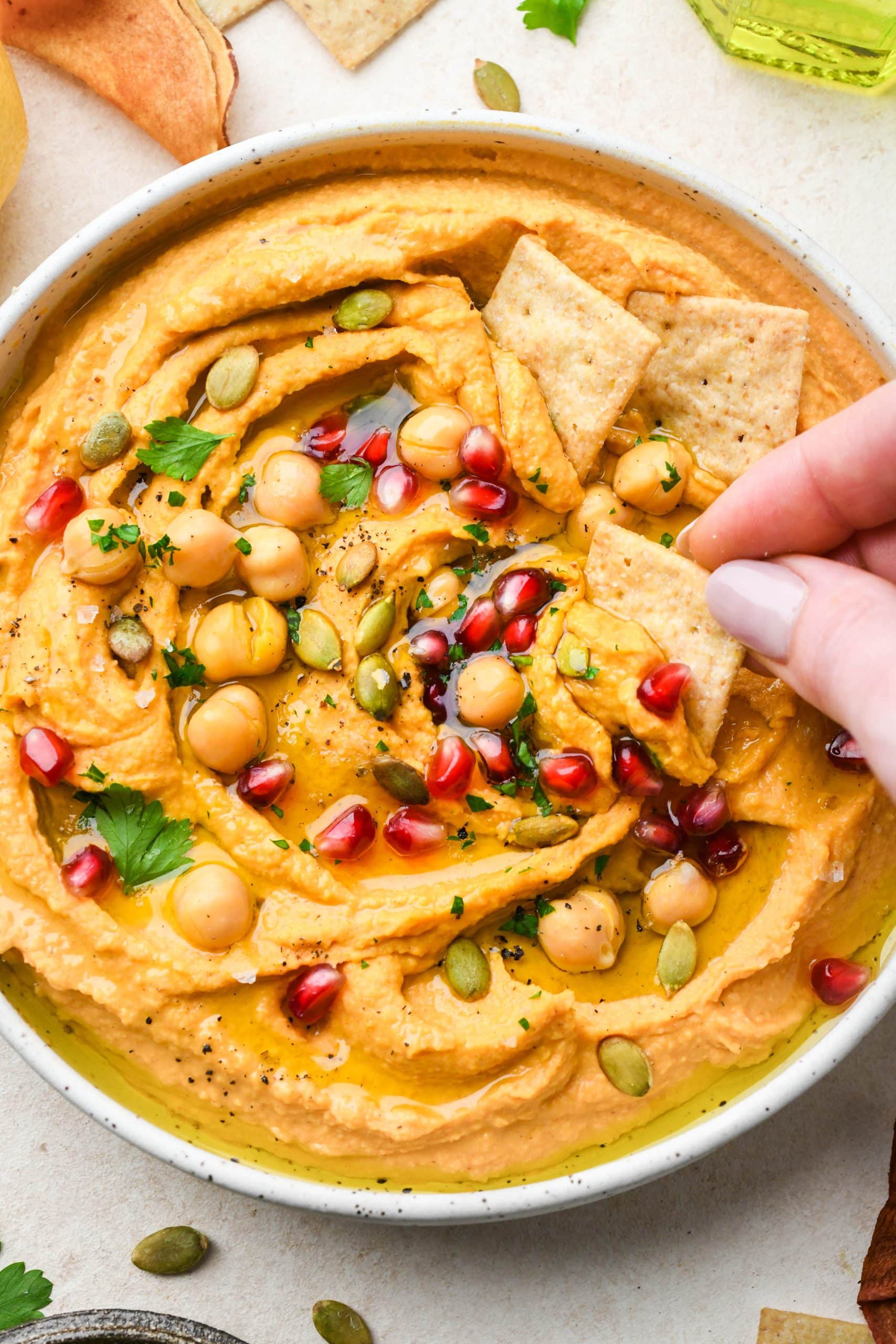 A shallow bowl of swirled sweet potato hummus topped with fresh herbs, pumpkin seeds, olive oil, and pomegranate with a hand dipping a small cracker into the dip.