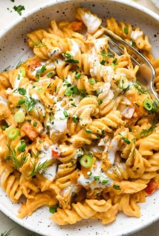 A shallow speckled bowl of creamy dairy free buffalo chicken pasta topped with fresh herbs, dairy free ranch, and a fork angled into the bowl.