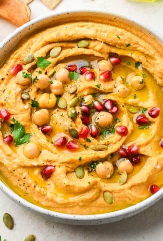 Close up image of a bowl of creamy sweet potato hummus topped with all the things - pomegranate, fresh parsley, olive oil, chickpeas, and flaky salt and pepper.