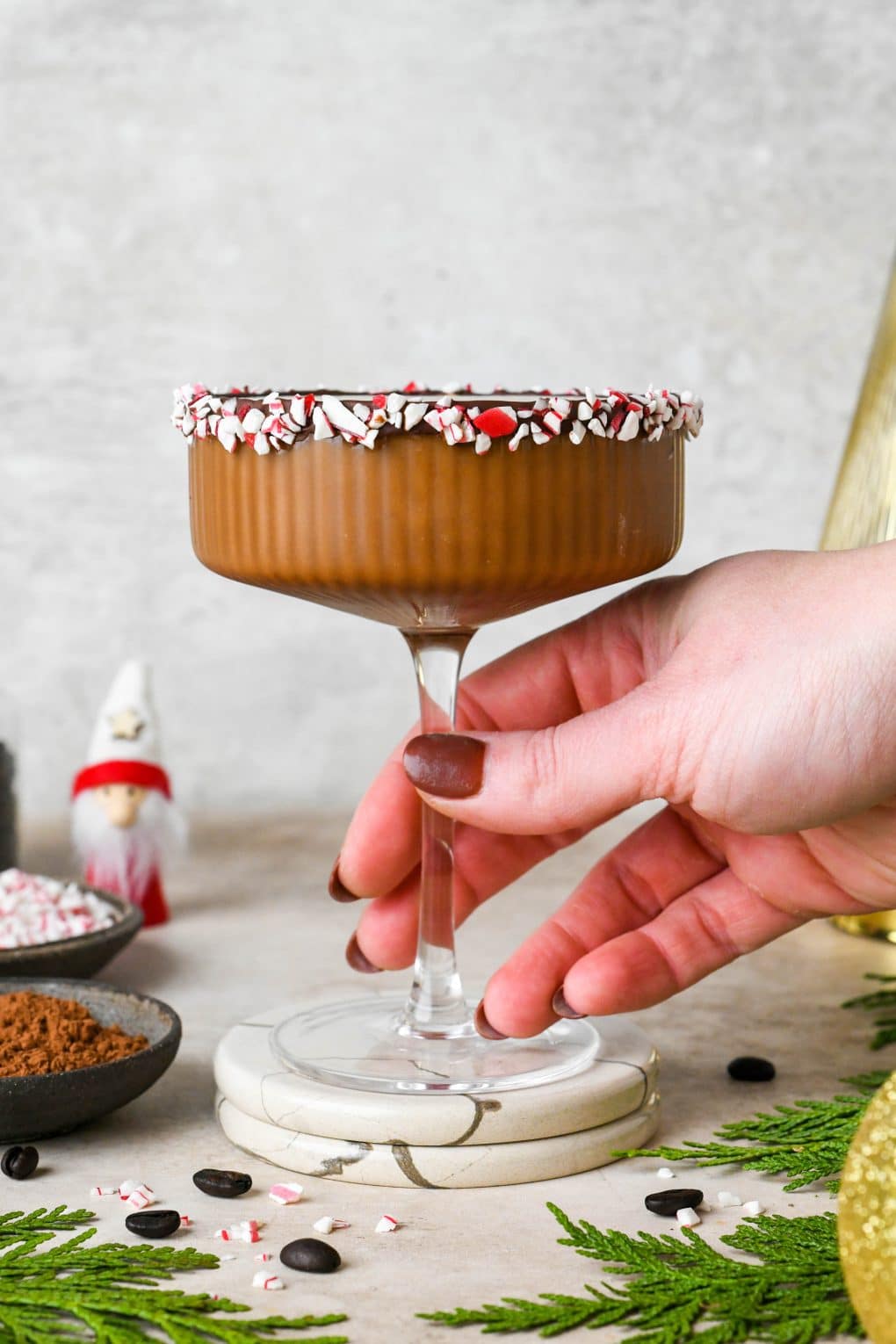 A peppermint mocha espresso martini in a martini glass with the rim lined with chocolate and peppermint candy, a hand reaching into the frame to hold the stem.