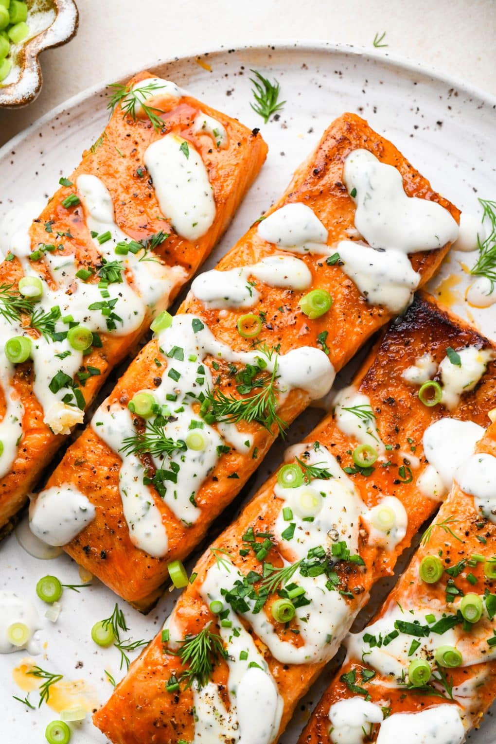 Four buffalo salmon filets on a white speckled plate baked to perfection and topped with a drizzle of dairy free ranch dressing, fresh herbs, and green onions.