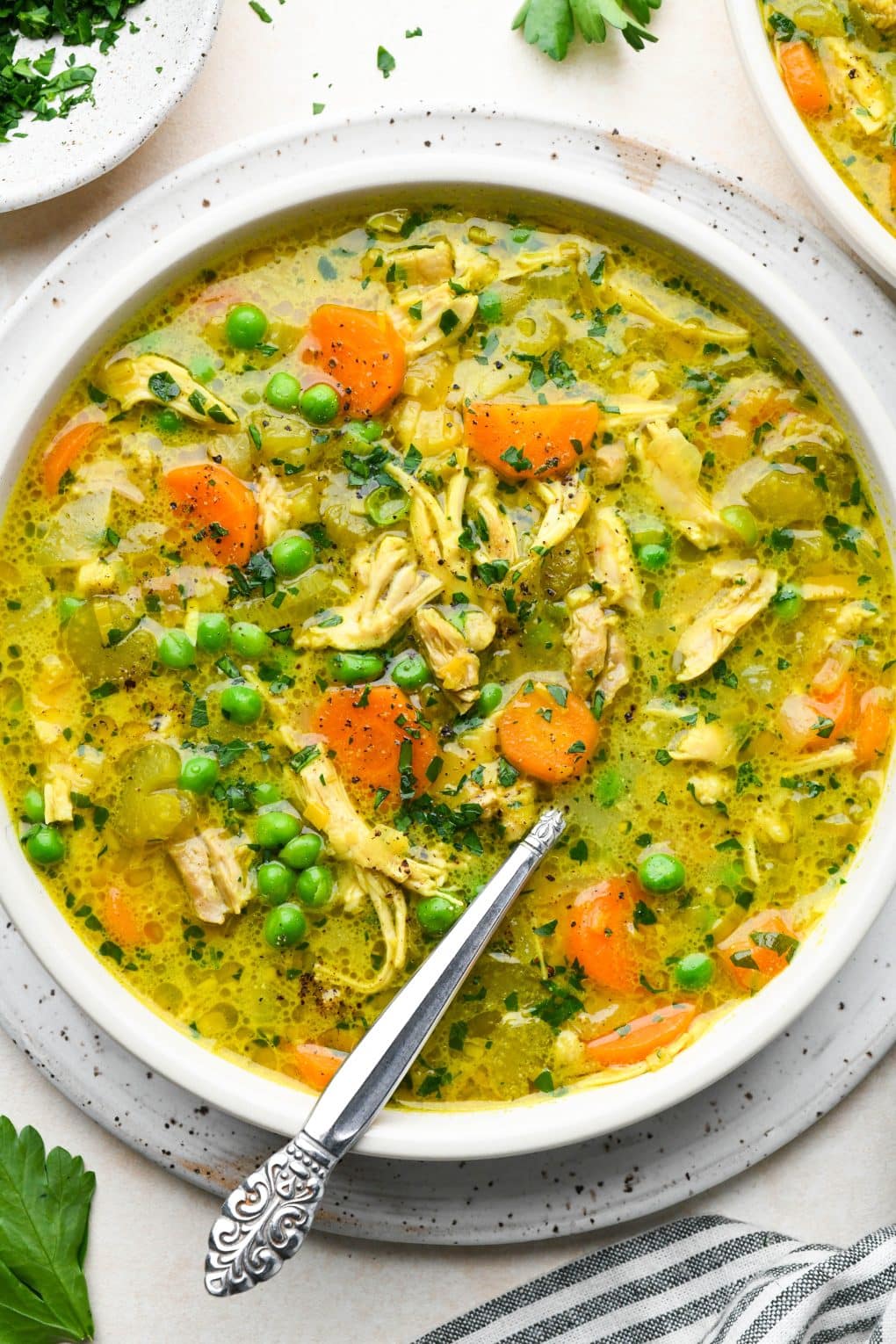 A large shallow bowl of bright yellow turmeric chicken soup with peas and fresh herbs. On a cream colored background.